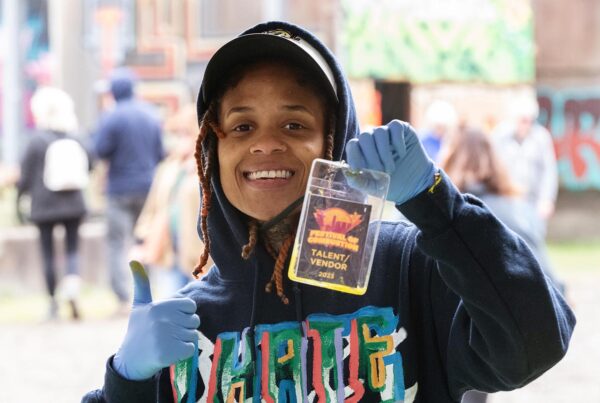 A youthful black woman with a big smile in a blue hoodie with colorful graphics holds up a " Vendor / Talent" ID badge and beams at the camera.