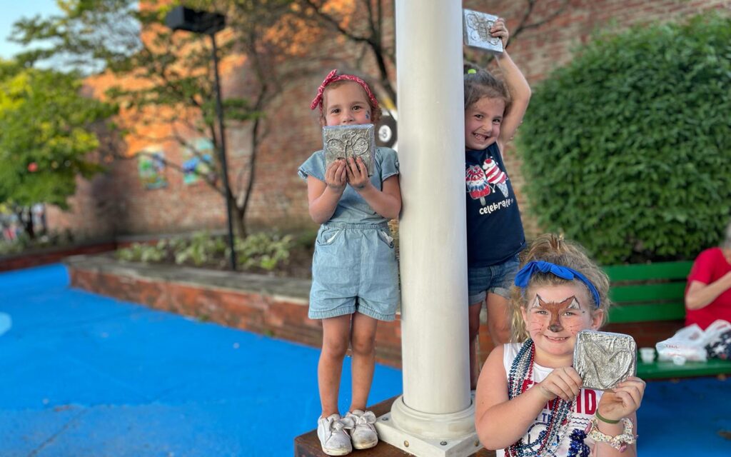 Three young white girls display their cast aluminum artworks as they stand on a newly-installed, artistically-designed light post. 