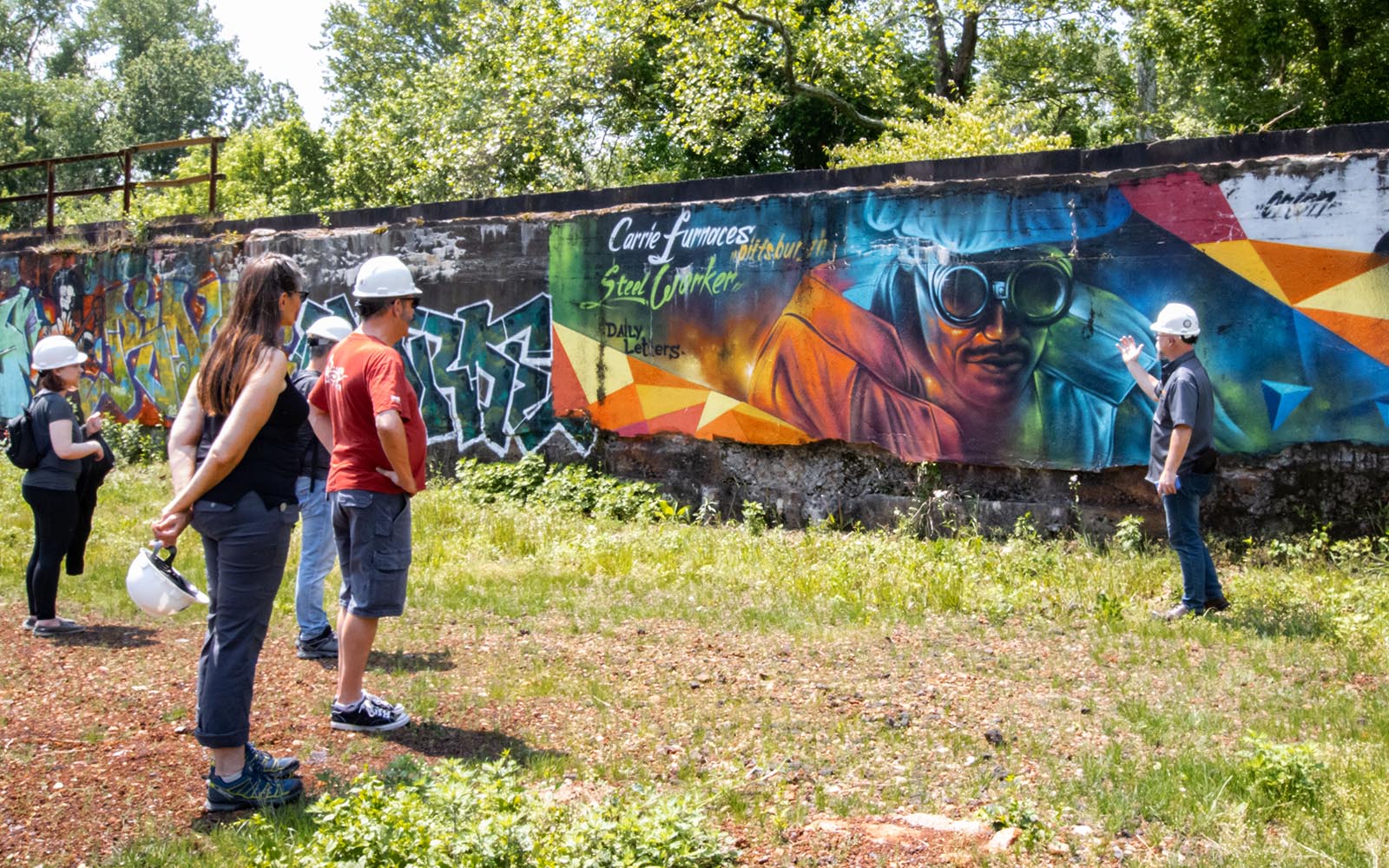 A tour guide gestures to a graffiti mural of a steelworker while guests view it.