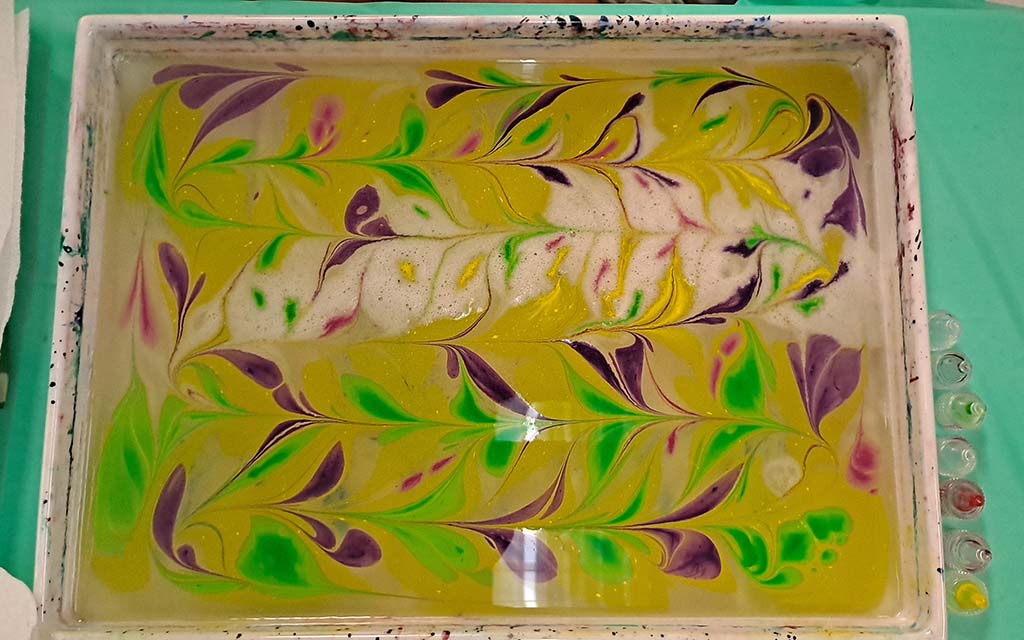 A tray is filled with water with ink dripped and stylized on the surface of the water. The background is yellow . The design looks like white and purple abstract flowers with green leaves.