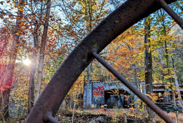 A rusting metal wheel crisscrosses the image revealing mosaic artwork in a fall landscape, viewable through its spokes.