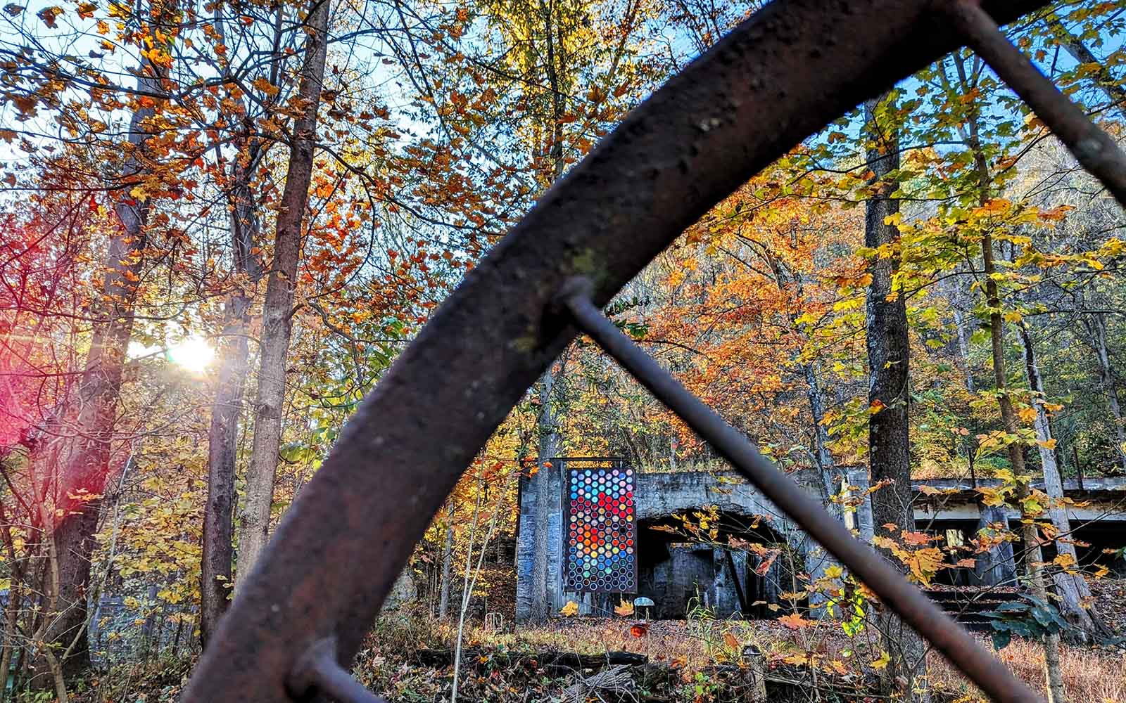 A rusting metal wheel crisscrosses the image revealing mosaic artwork in a fall landscape, viewable through its spokes.
