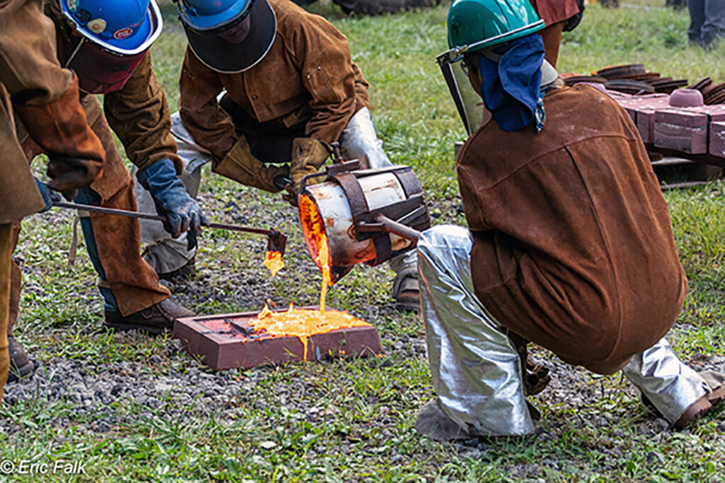 Students work together to pour molten iron from a ladle into a sand mold.