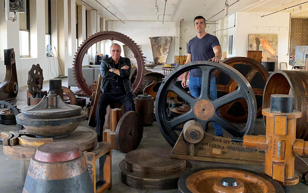 Two men stand in a room surrounds by wooden wheels and other industrial foundry patterns.