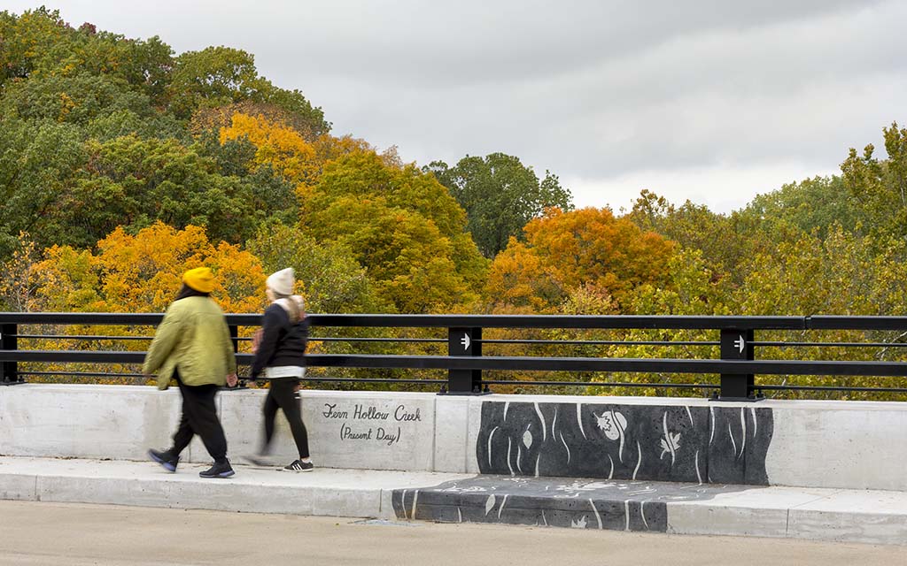 Two pedestrians cross a bridge with fall trees in the background and art embedded in the sidewalk.