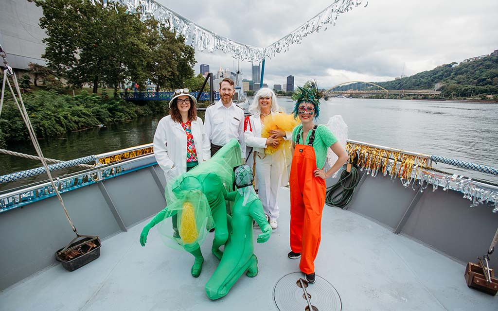 Five people in costume pose with a boat captain on the bow of a vessel with a river in the background.