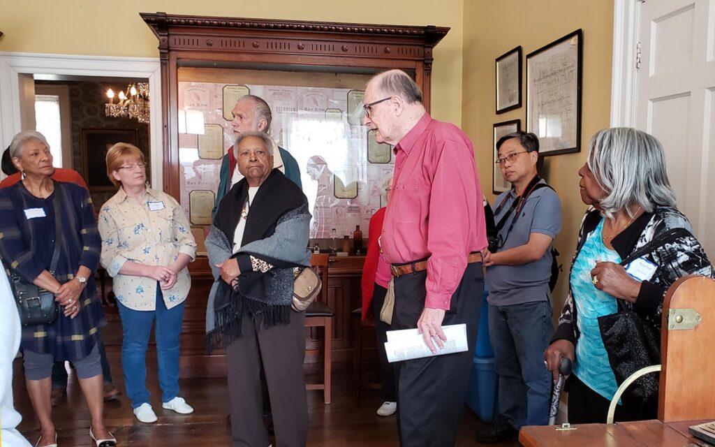 A multi-ethnic group of elders gathers around a docent in a historic home.