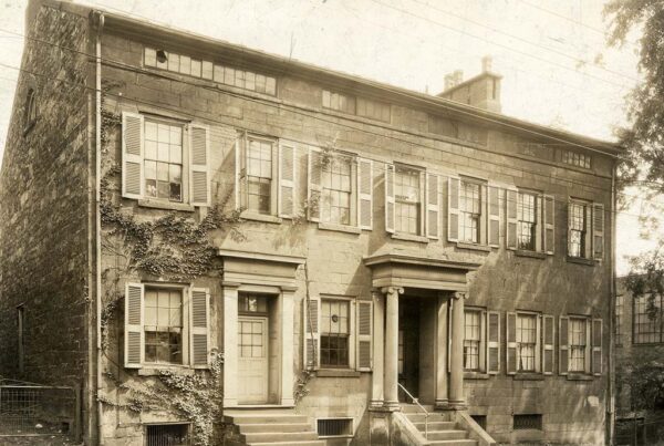 A sepia-toned image of a large colonial style home with a front entrance with columns that offers steps down to a sidewalk that runs right along the street. A second entrance to the right is less ornate and disrupts the home's symmetry.