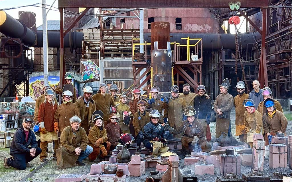 A group photo of 27 people in leathers, hard hats and uplifted masks posed on the pour floor with mold in front of them and the furnace behind them.