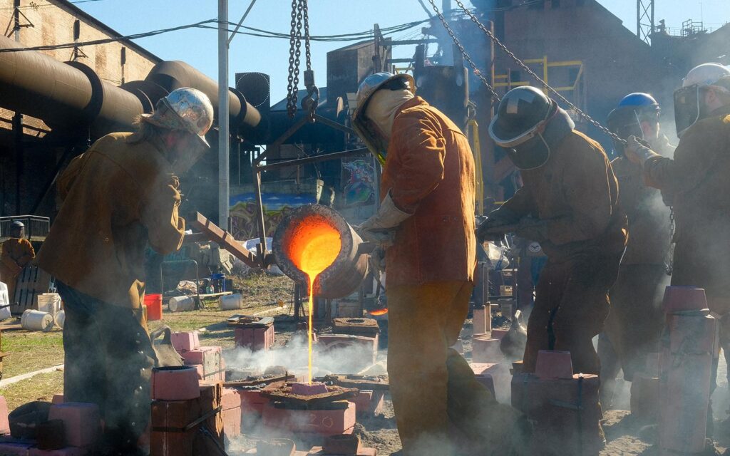 Five people in protective gear pour iron from a large ladle into resin-bonded sand molds.