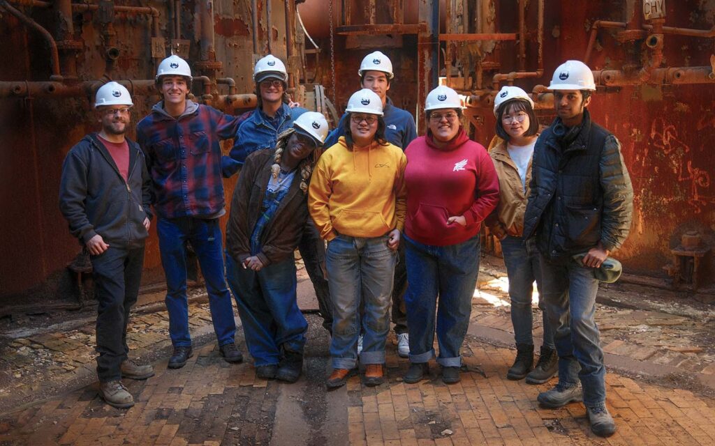 A group of nine people in hard hats pose for a photo in an industrial setting.