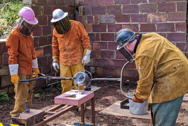 Three metal workers in leather protective gear pour molten aluminum into a mold.