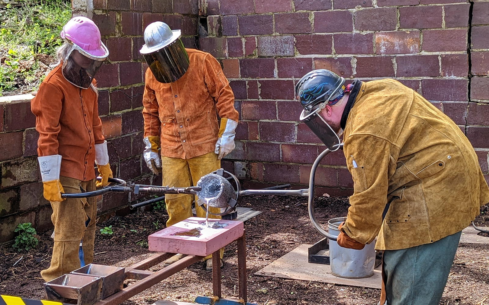 Three metal workers in leather protective gear pour molten aluminum into a mold.