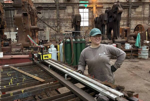A white woman in a long sleeved t and ball cap poses for the camera in an industrial space.
