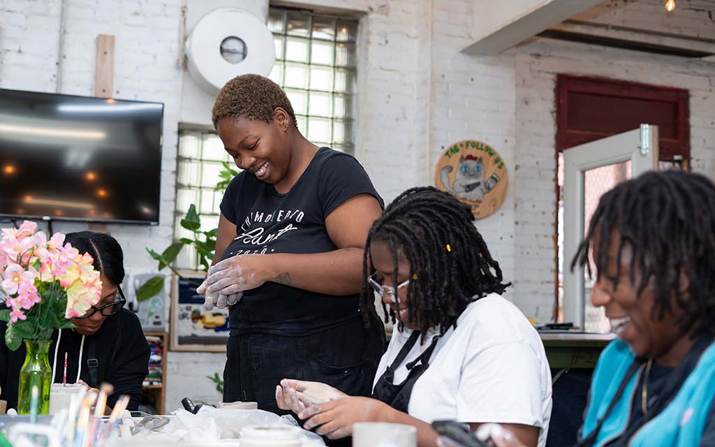 Black women sit at a table with flowers in a vase and appear to be working on baking something as their hands are covered in flour.