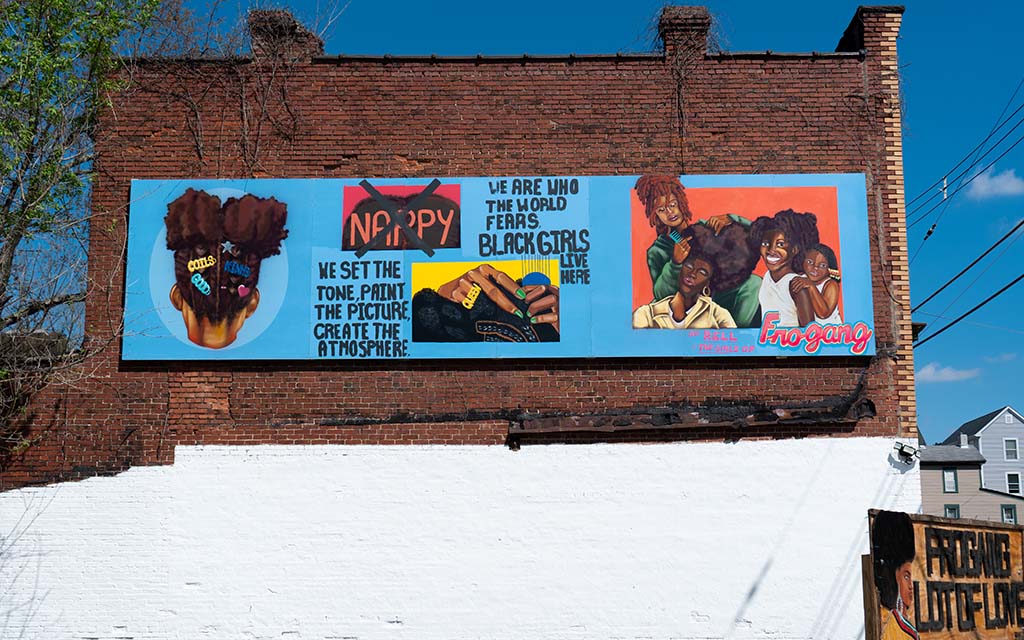 A mural on panels affixed to a brick building with a blue background that features representations of black women with natural hair and inspirational quotes.