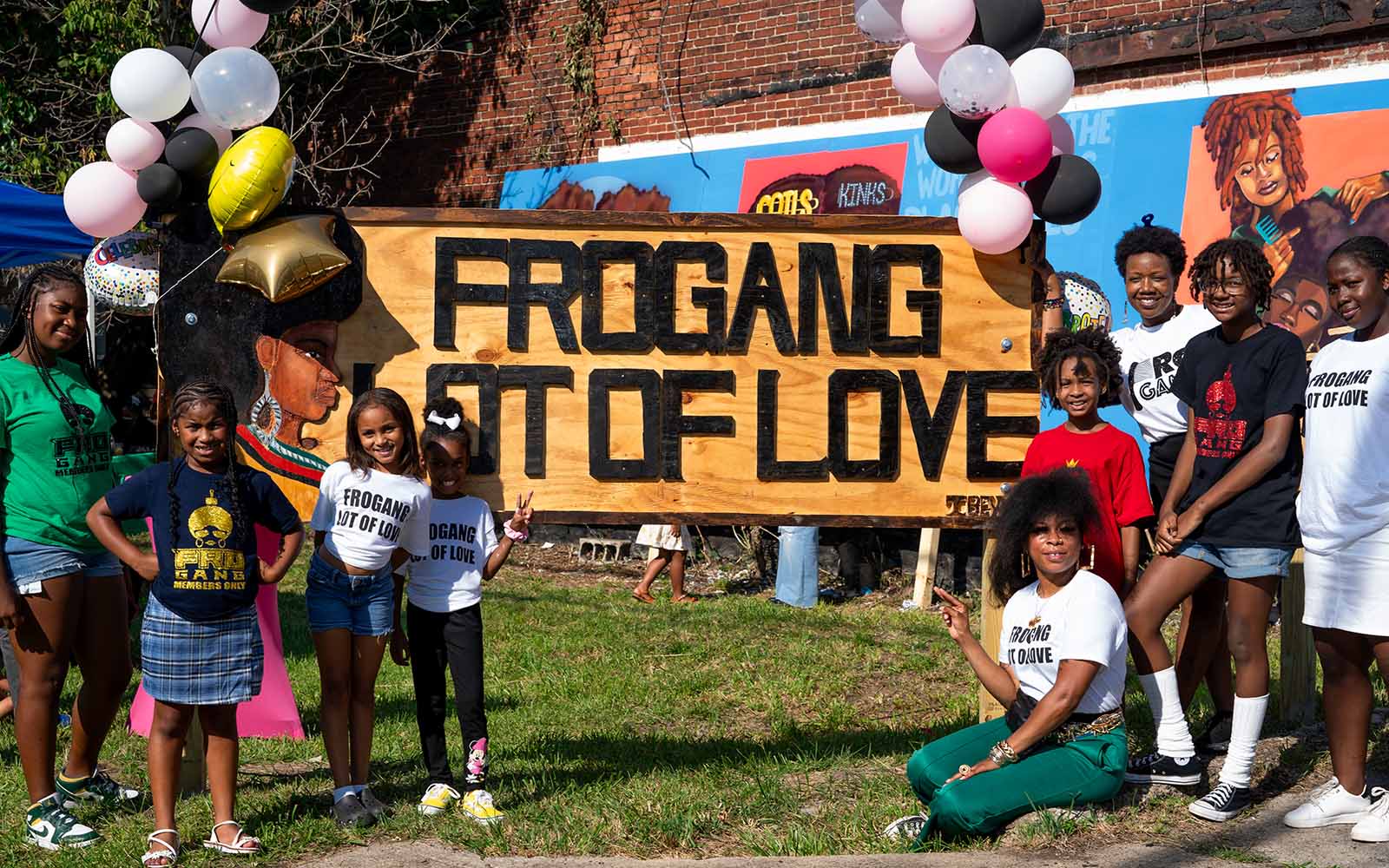 A group of young black women of a variety of ages with natural hair pose with a sign that reads "FROGANG LOT OF LOVE" with balloons on it.