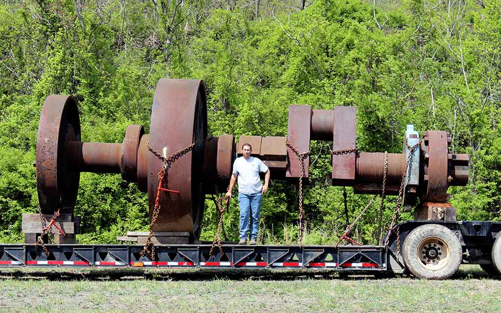 A man stands on a flatbed trailer in front of a crankshaft that nearly doubles his height that could be 30 feet long.