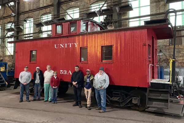 Seven people stand in front of a red caboose inside a large industrial space.