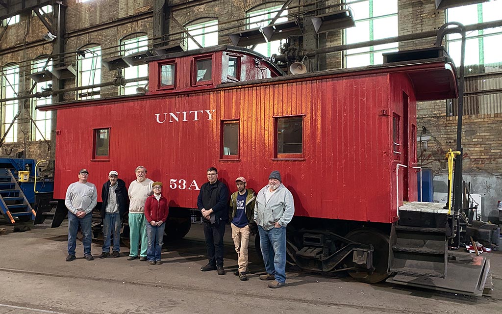 Seven people stand in front of a red caboose inside a large industrial space.