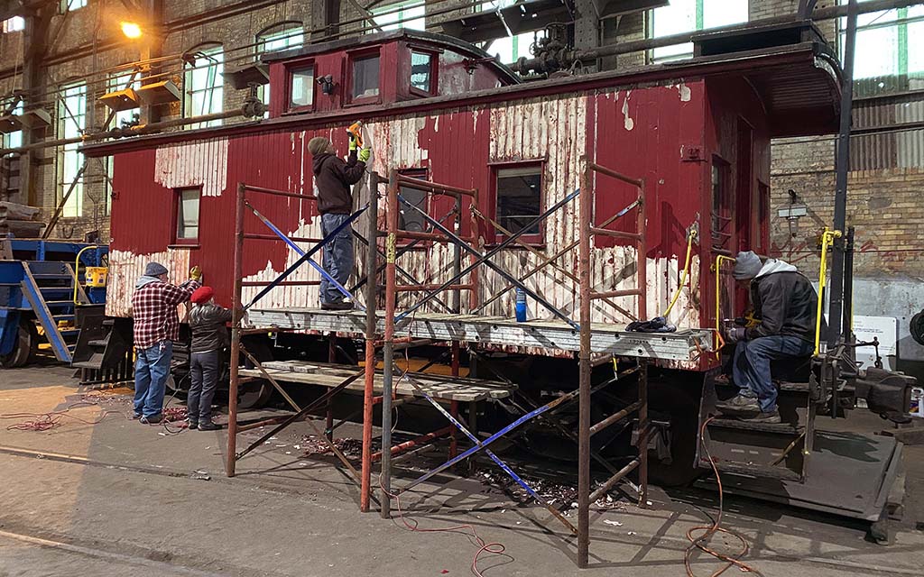 Four people, two on the ground and two on scaffolding, strip paint from an old caboose.