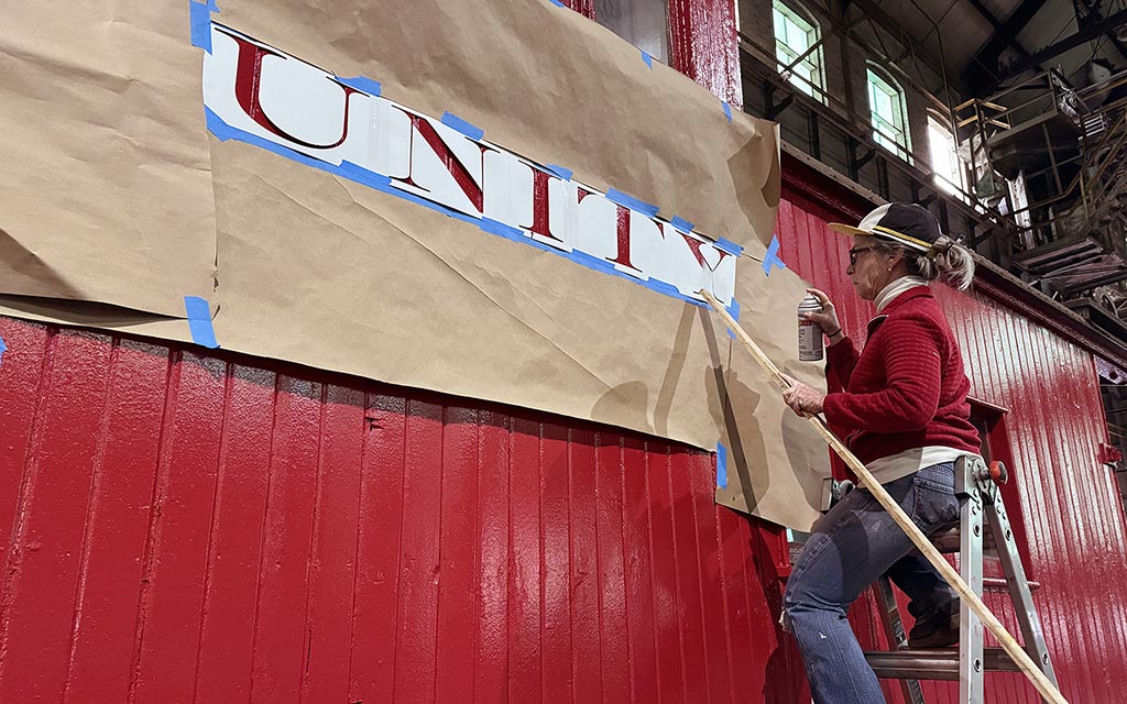 A woman on a ladder lifts her arm to paint the word "Unity" which is masked off by paper. 