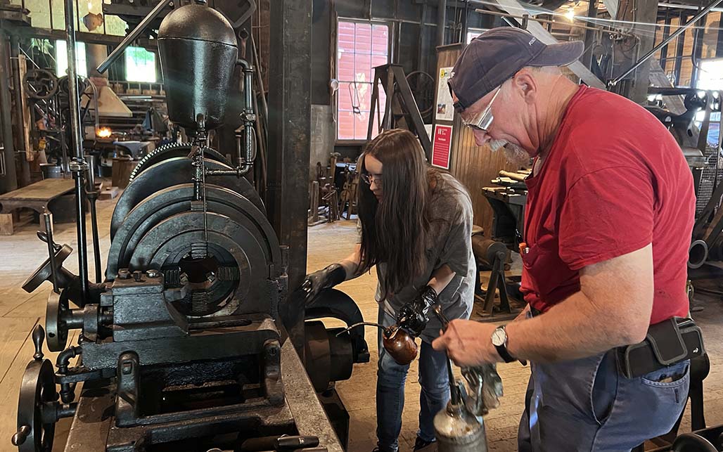 A young woman with long brown hair works on a machine with an older gentleman wearing goggles.