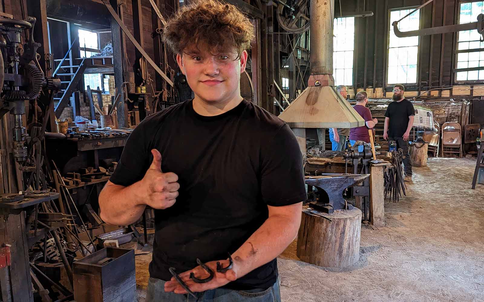 A young broad-shouldered man with a mop of curly hair gives a thumbs up with one hand and displays forged steelwork with the other.