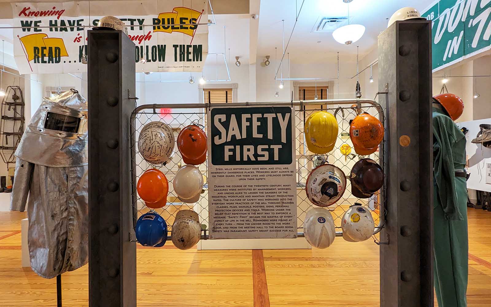 A sign that reads "Safety First" is flanked by a dozen hard hats hung on aluminum fencing.