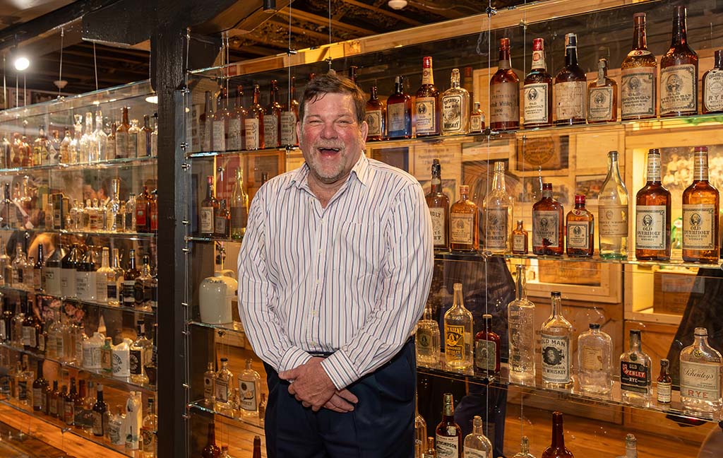 A jovial looking man with brown hair and a gray beard in a button down shirt and slacks gives a large smile in front of a wall of shelves filled with bottles. 