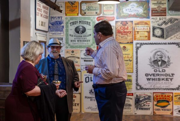 Three people in conversation in a gallery with old booze labels and ads reproduced on a wall behind them.
