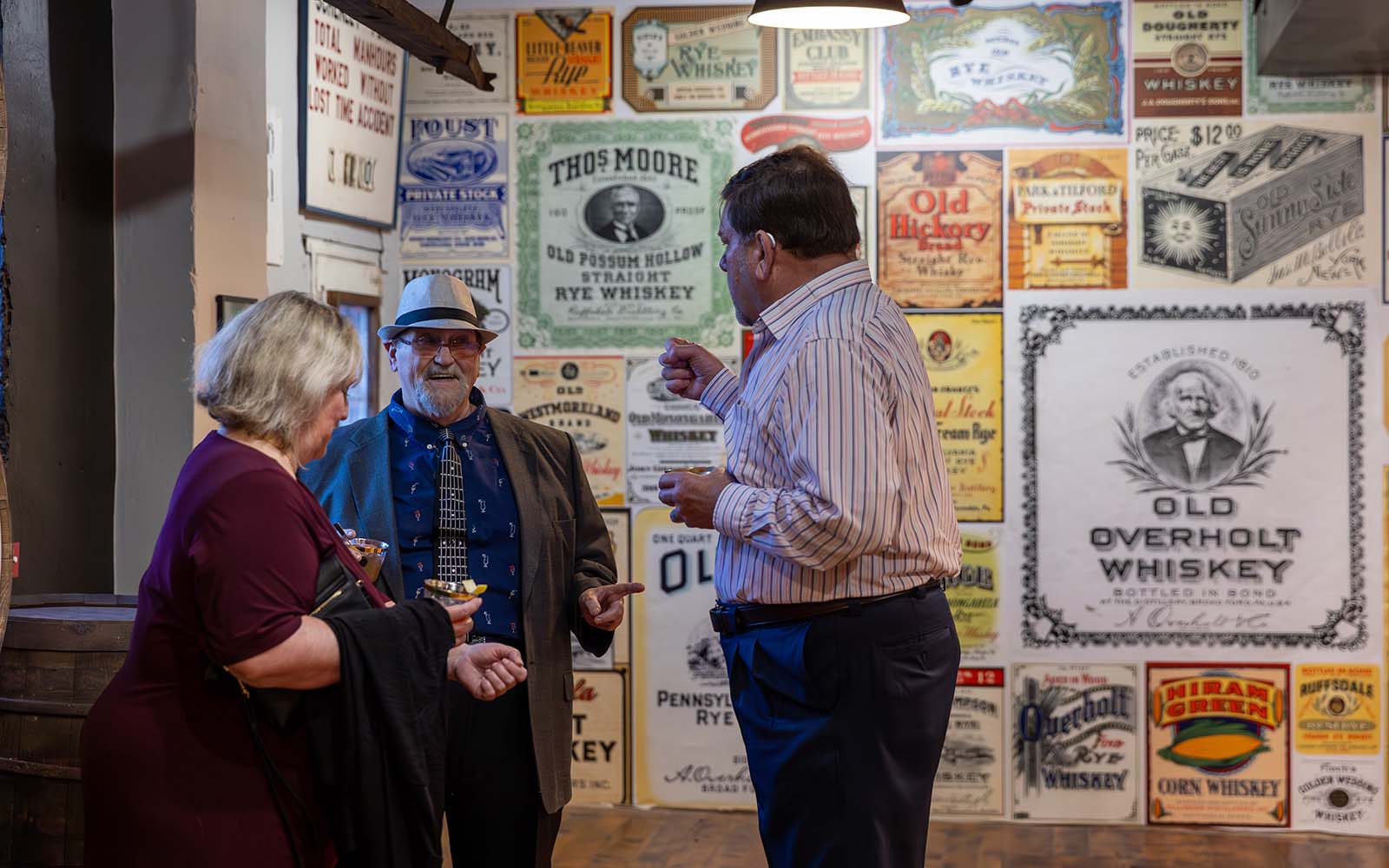 Three people in conversation in a gallery with old booze labels and ads reproduced on a wall behind them.