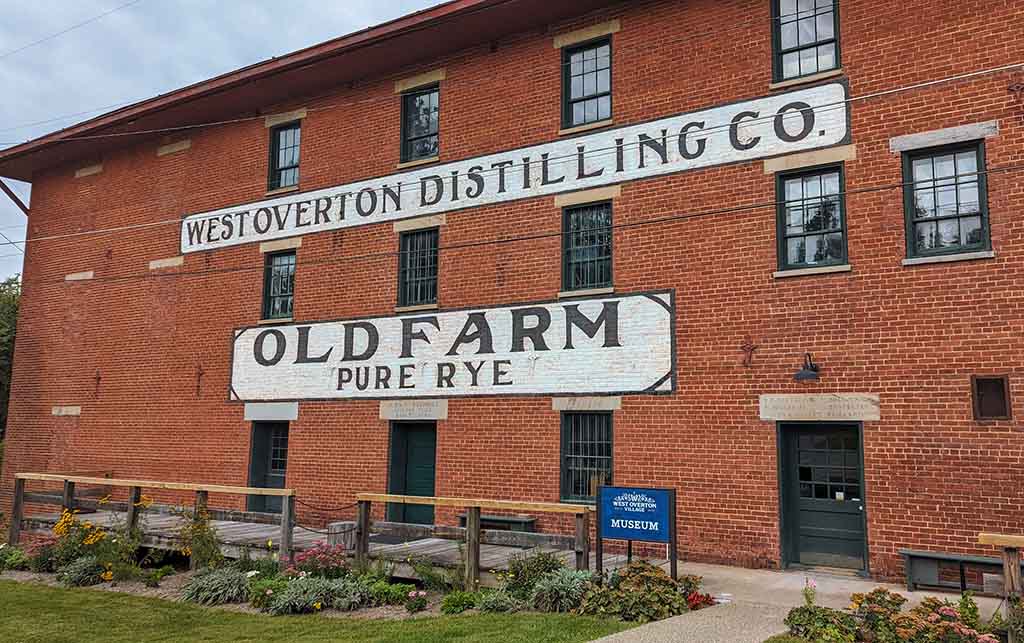 A large brick building with "West Overton Distiling Co." painted on it, along with "Old Farm Pure Rye. A small sign reads "Museum" near an entrance.