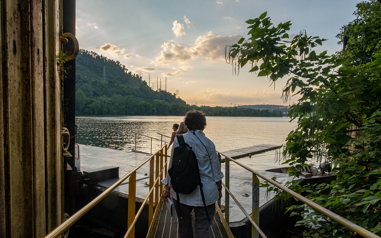 An over the shoulder shot of several people caring notebooks in their hands, walking on a gangway towards a dock on a river with a setting sun.