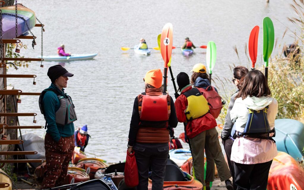 Kayakers line up by the river with paddles. 