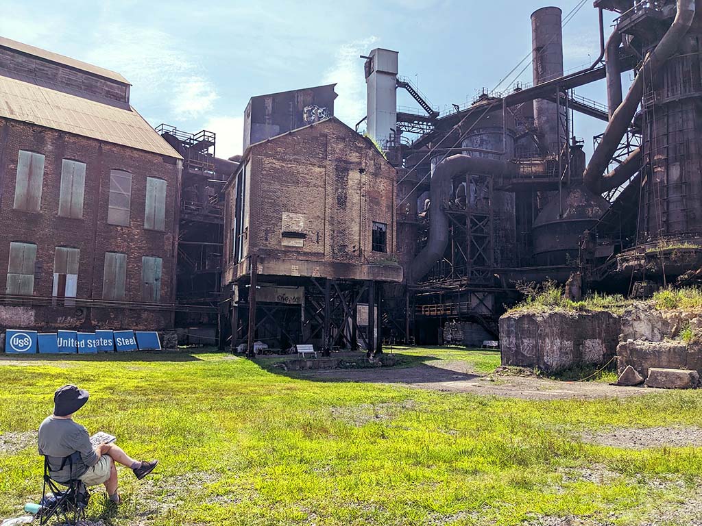 A figure sits in a camp chair with a sketchbook looking up at Furnace #7.