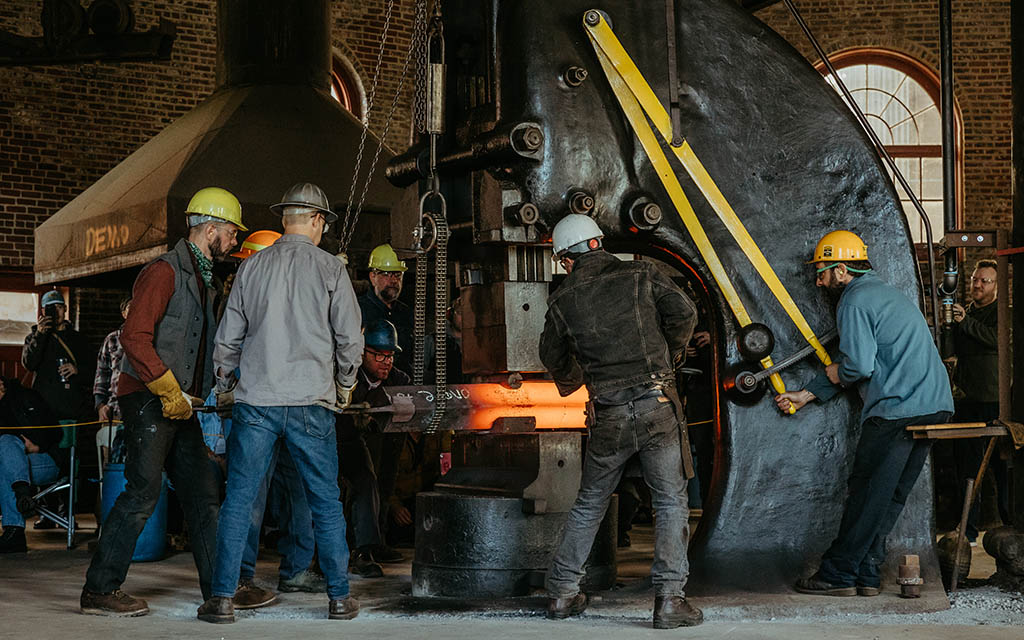 Six men in hard hats and safety gear work a large power hammer.