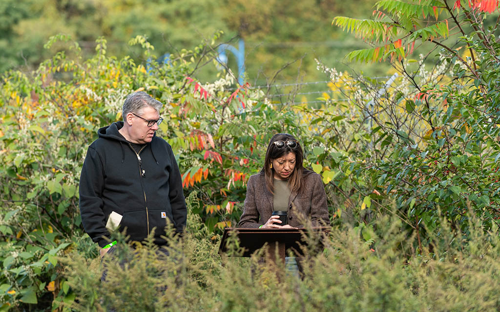 A white man in a black hoodie and a white woman in a fall blazer look down at the cast iron plaques in a garden space.