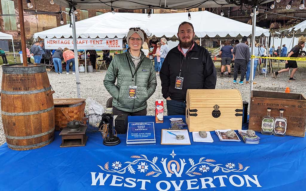 A man and woman stand behind a table with historical objects and a tablecloth that reads West Overton.