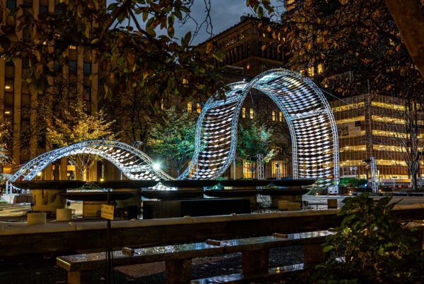 A wavelike structure is lit in blue lights and see in a park at dusk.