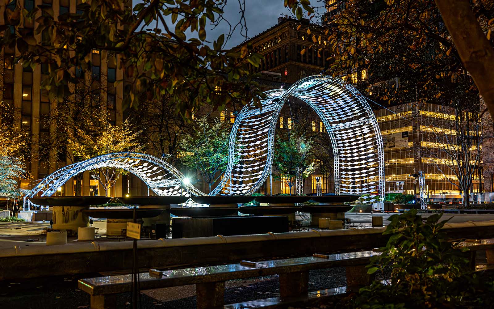 A wavelike structure is lit in blue lights and see in a park at dusk.