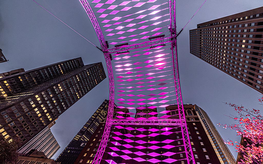 See from below and lit by purple light, the art installation looks like a bridge across the sky with skyscrapers surrounding it creating a vanishing point.