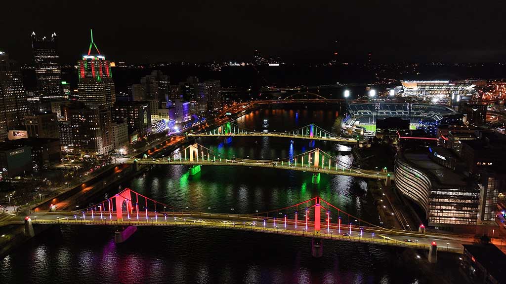 A night aerial view of three bridges lit in various colors.