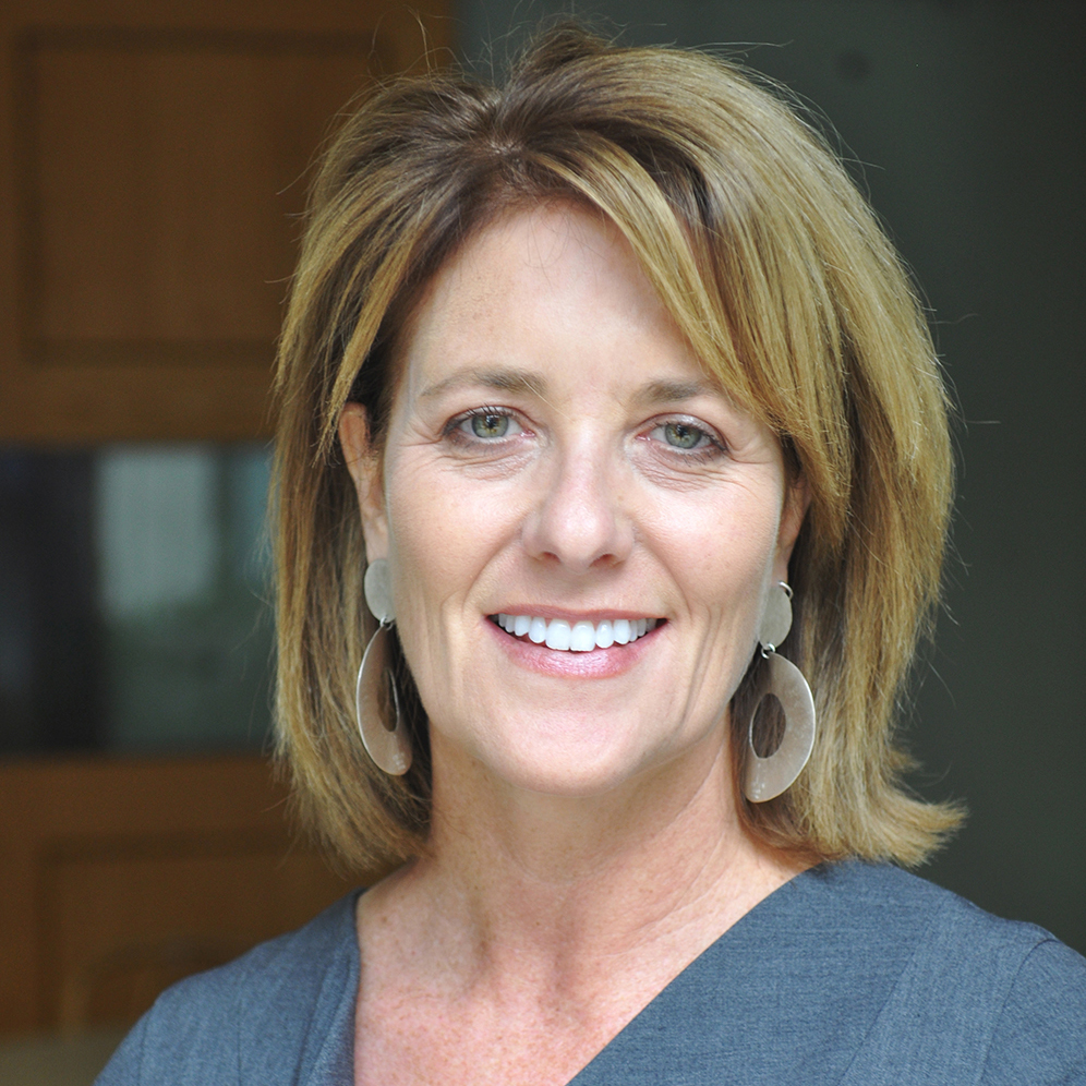 A white woman in a professional dress with large earrings, sandy blonde hair, and a nice smile.
